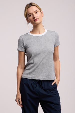 A person wears the MILEY TEE by Velvet by Graham & Spencer, a grey cropped ribbed crewneck top, paired with navy blue pants, standing against a plain light background.