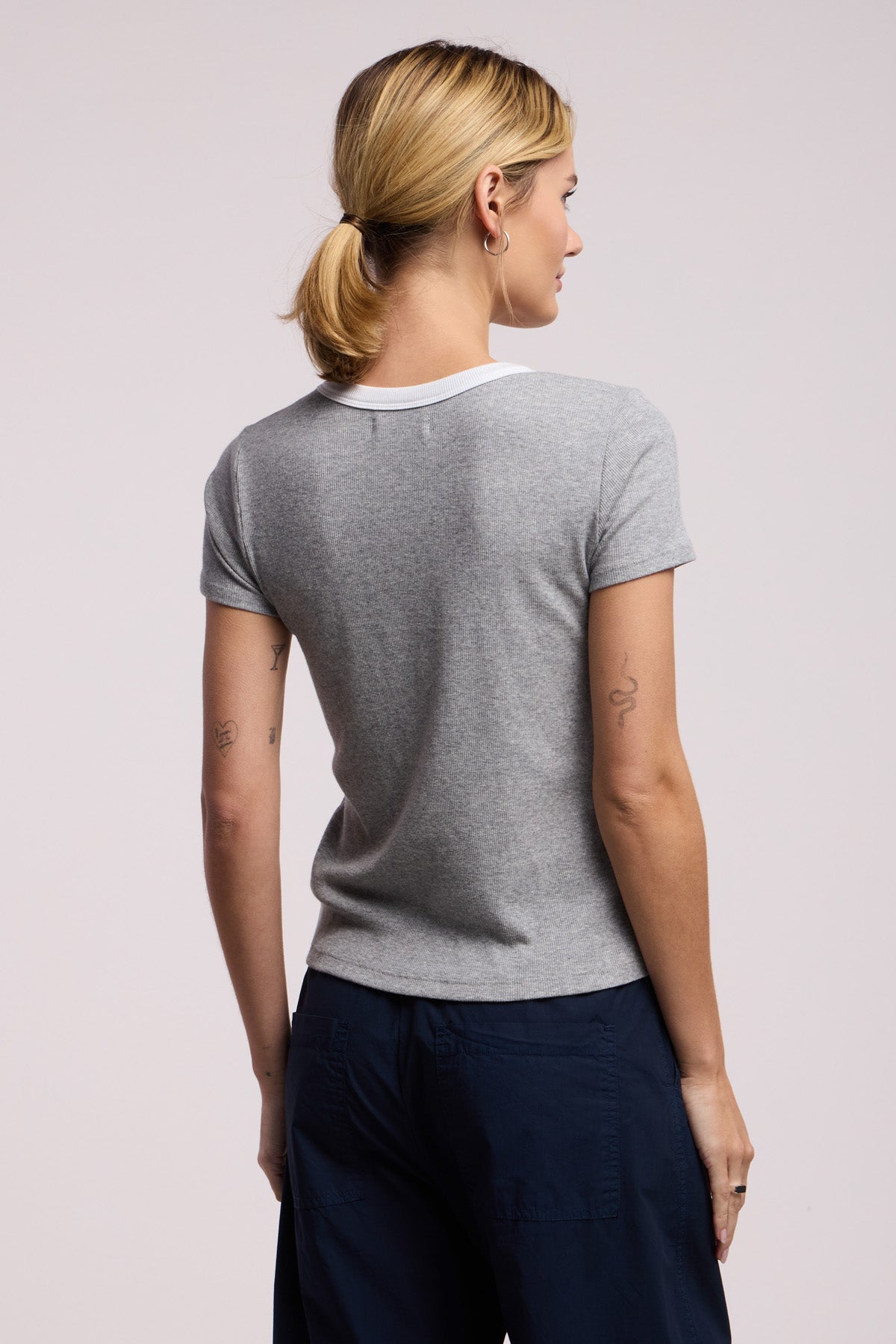 A person with blonde hair in a low ponytail wears the MILEY TEE by Velvet by Graham & Spencer—a gray cropped ribbed crewneck top—paired with dark pants, shown from the back against a light background.-41588377256129