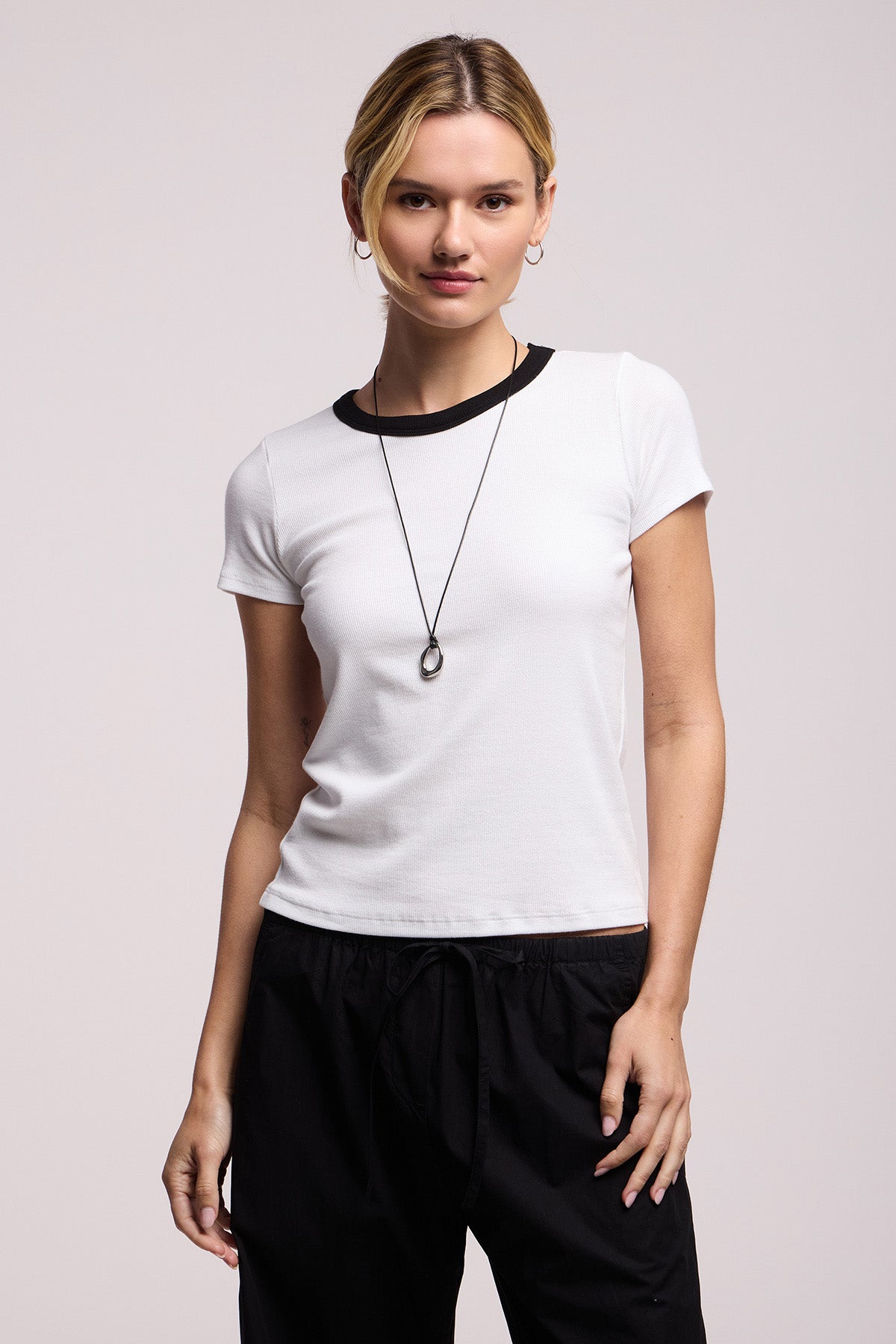 A person wears the MILEY TEE by Velvet by Graham & Spencer—a white short-sleeve fitted t-shirt with a black collar—paired with black pants and a long necklace, standing against a plain background.