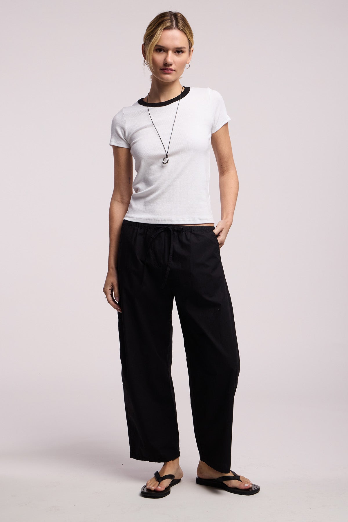 A person stands against a plain background wearing the Velvet by Graham & Spencer MILEY TEE, a white cropped ribbed top with a fitted silhouette, paired with black drawstring pants, black flip-flops, and a necklace.