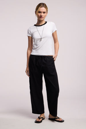 A person stands against a plain background wearing the Velvet by Graham & Spencer MILEY TEE, a white cropped ribbed top with a fitted silhouette, paired with black drawstring pants, black flip-flops, and a necklace.