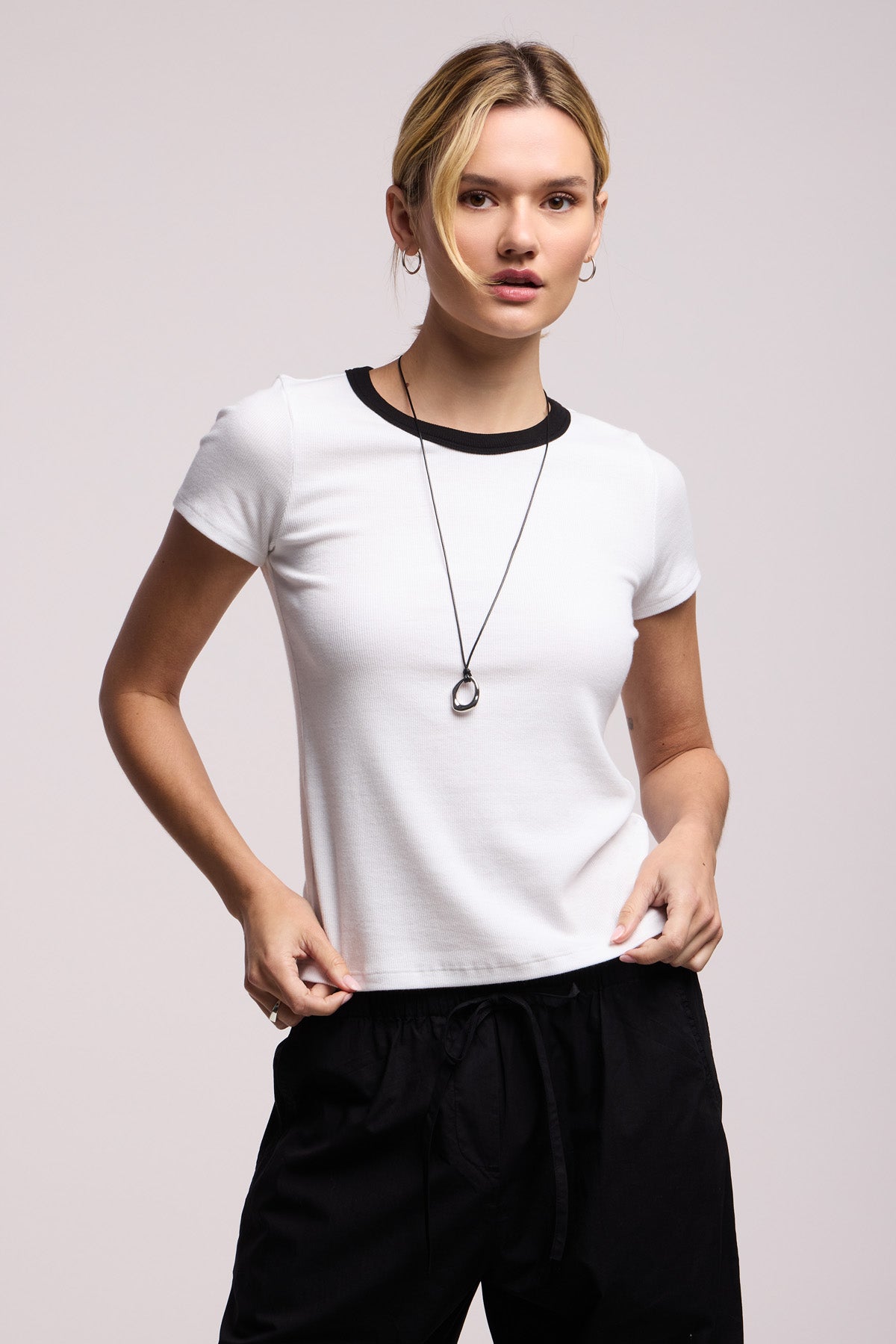 Wearing the MILEY TEE by Velvet by Graham & Spencer—a white cropped ribbed tee with black trim—styled with black pants and a necklace, standing against a plain background.