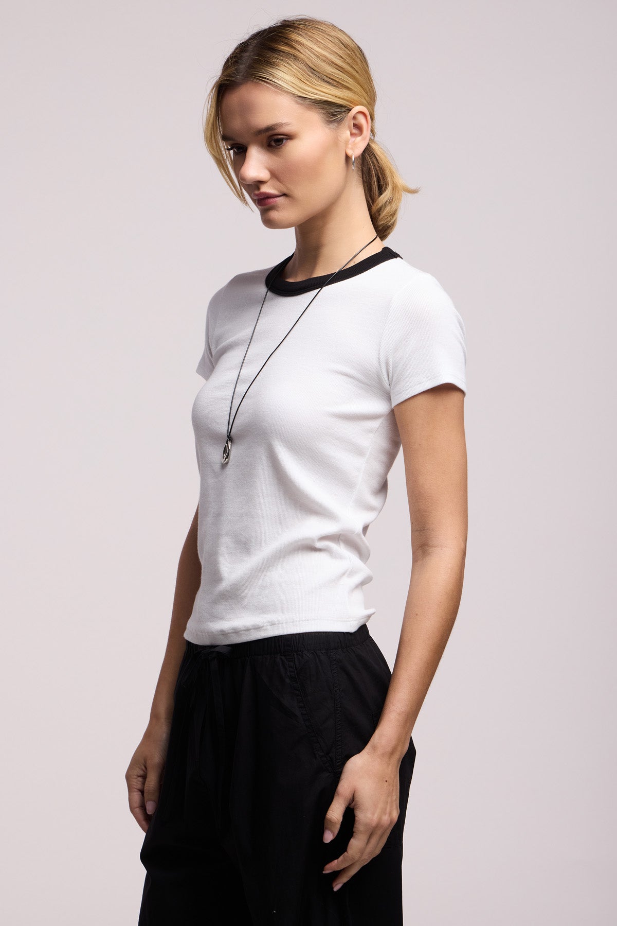 Woman in profile wears the Velvet by Graham & Spencer MILEY TEE, a fitted cotton-modal blend white short-sleeve t-shirt with a black collar, paired with black pants and a necklace, against a plain background.
