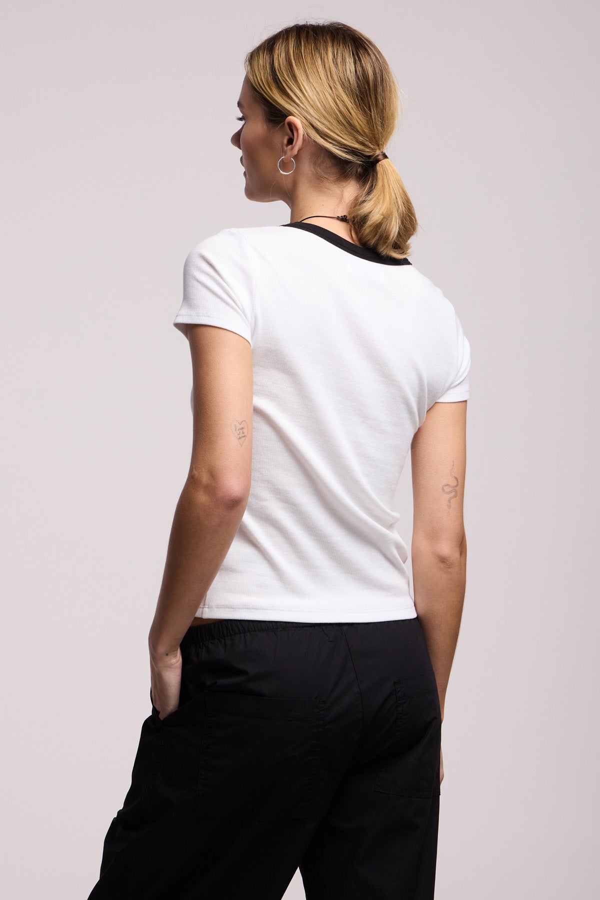 A person with blonde hair in a low ponytail is shown from the back wearing the MILEY TEE by Velvet by Graham & Spencer, a white cotton-modal blend short-sleeve shirt with a black collar, paired with black pants.