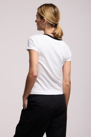 A person with blonde hair in a low ponytail is shown from the back wearing the MILEY TEE by Velvet by Graham & Spencer, a white cotton-modal blend short-sleeve shirt with a black collar, paired with black pants.