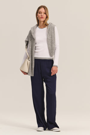 A woman stands facing forward, wearing the NOVATO TEE by Velvet by Graham & Spencer and navy wide-leg pants, with a gray sweater draped over her shoulders. She accessorizes with a light bag and white shoes.