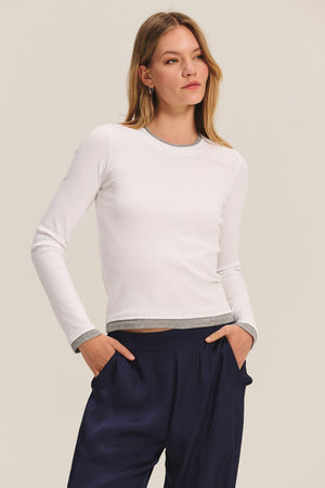A woman models the NOVATO TEE by Velvet by Graham & Spencer—a ribbed modal top with gray trim and a double-layered look—paired with navy blue pants, standing with hands in her pockets against a plain background.