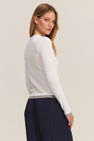 A woman with long hair models the NOVATO TEE by Velvet by Graham & Spencer—a white ribbed modal top with gray trim—paired with navy pants, standing at a slight angle to the camera.