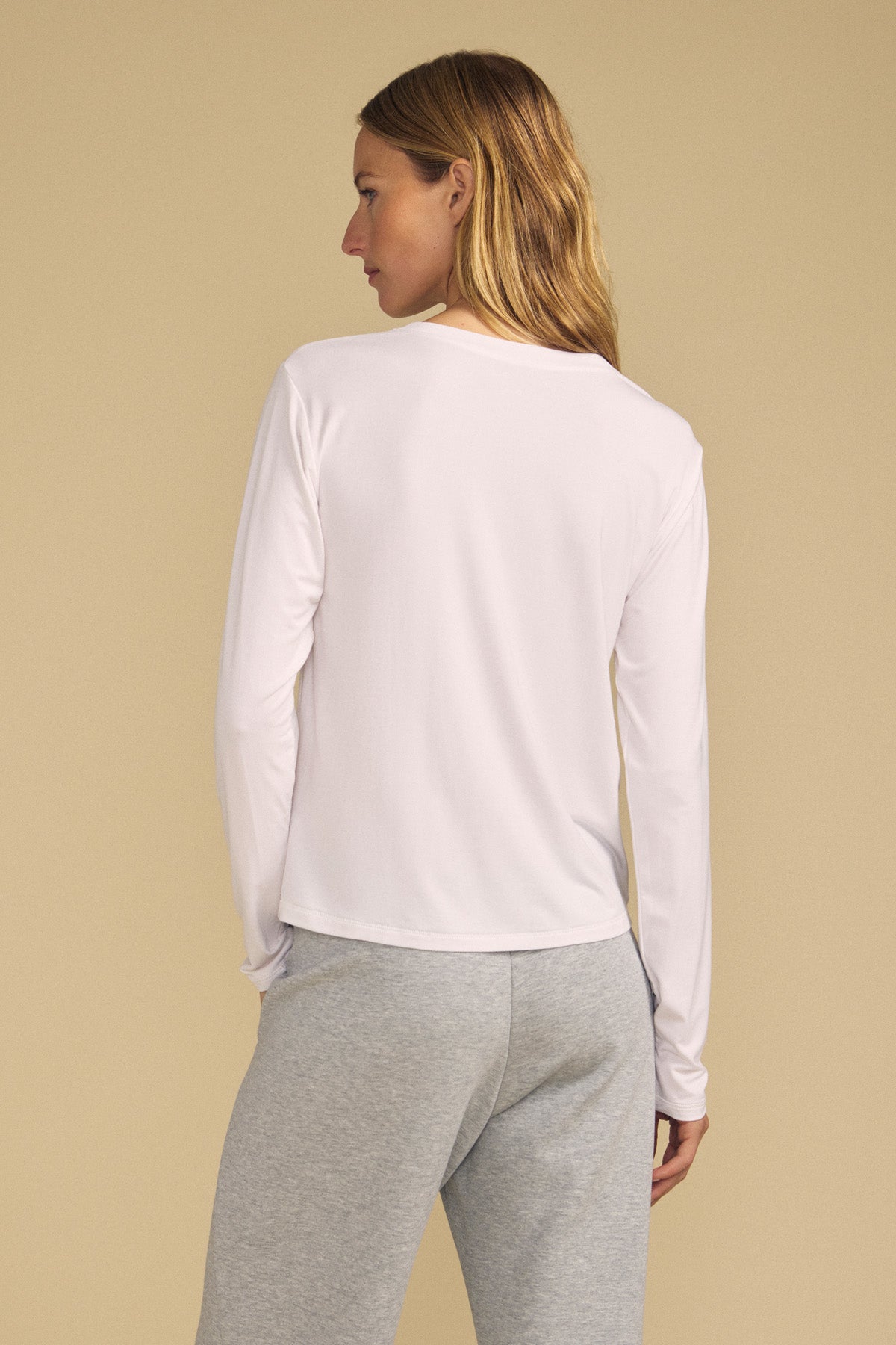 A person stands with their back to the camera, wearing the long-sleeve BARBARA TEE by Velvet by Graham & Spencer in white, paired with light grey pants, set against a beige background.-41814380773569
