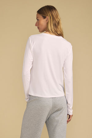 A person stands with their back to the camera, wearing the long-sleeve BARBARA TEE by Velvet by Graham & Spencer in white, paired with light grey pants, set against a beige background.
