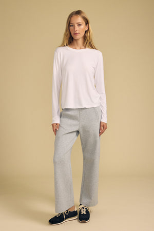 A woman stands against a beige background wearing the Velvet by Graham & Spencer BARBARA TEE, paired with gray wide-leg pants and black sneakers.