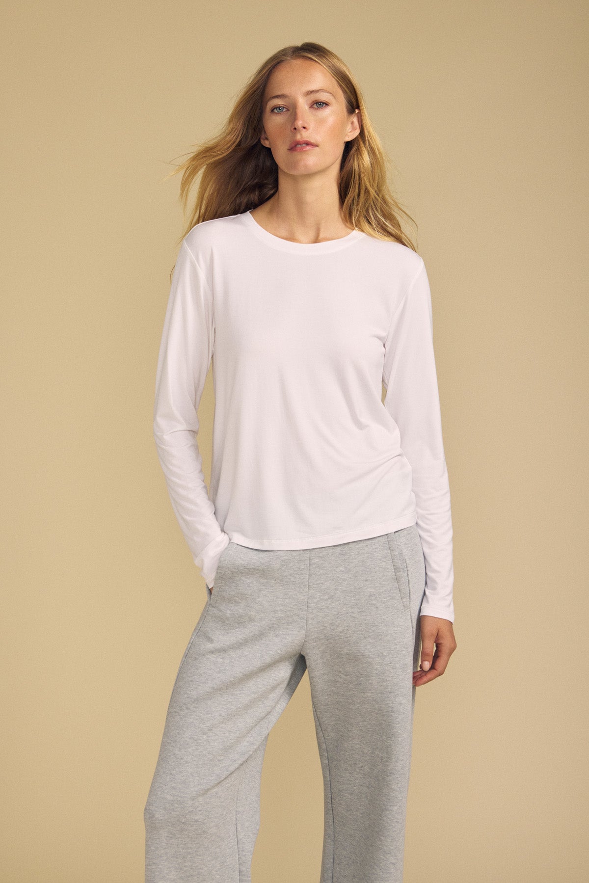 A woman stands against a beige background wearing the BARBARA TEE by Velvet by Graham & Spencer, paired with gray sweatpants and one hand in her pocket.