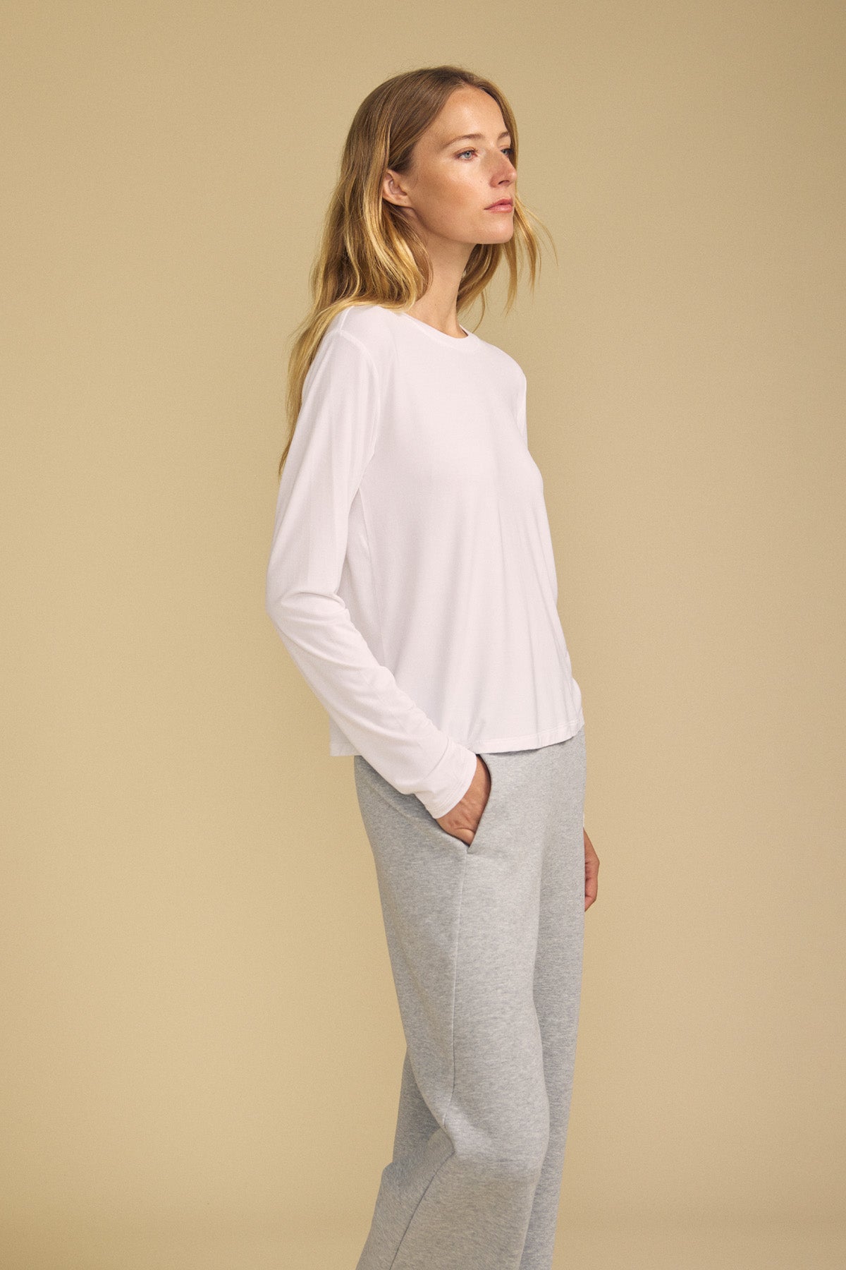 A woman stands sideways with her hands in her pockets, wearing the BARBARA TEE by Velvet by Graham & Spencer—a long-sleeve white top—paired with light gray sweatpants, set against a plain beige background.-41814380806337