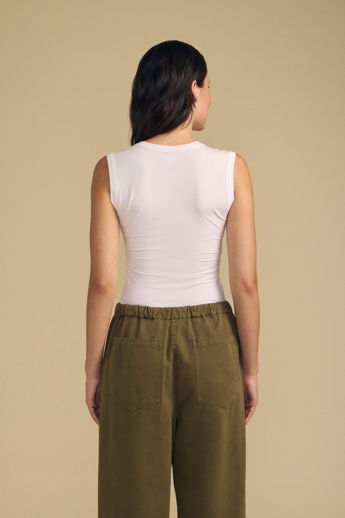 A person stands with their back to the camera, wearing the IYLA TANK TOP by Velvet by Graham & Spencer in white and paired with olive green pants, set against a plain beige background.