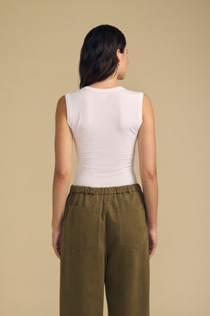 A person stands with their back to the camera, wearing the IYLA TANK TOP by Velvet by Graham & Spencer in white and paired with olive green pants, set against a plain beige background.