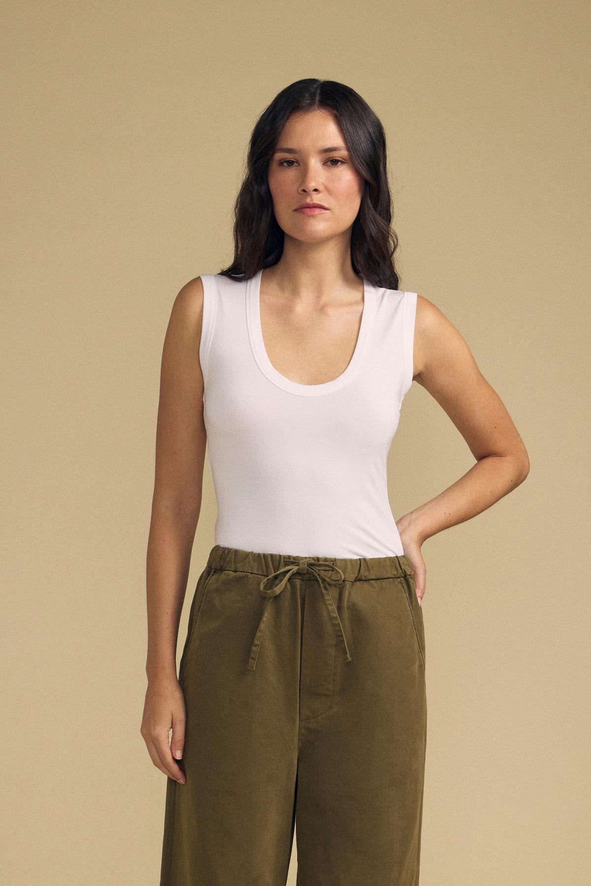 A woman with long dark hair stands facing forward, wearing the Velvet by Graham & Spencer IYLA TANK TOP in white and olive green drawstring pants against a plain beige background.