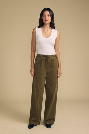 A woman stands facing forward against a plain beige background, wearing the IYLA TANK TOP by Velvet by Graham & Spencer—a white sleeveless top—paired with olive green wide-leg pants featuring a drawstring waist and black shoes.