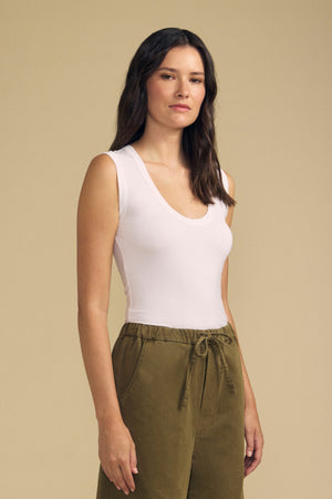 A woman with long dark hair wears the Velvet by Graham & Spencer IYLA TANK TOP in white and olive green high-waisted pants, standing against a plain beige background.