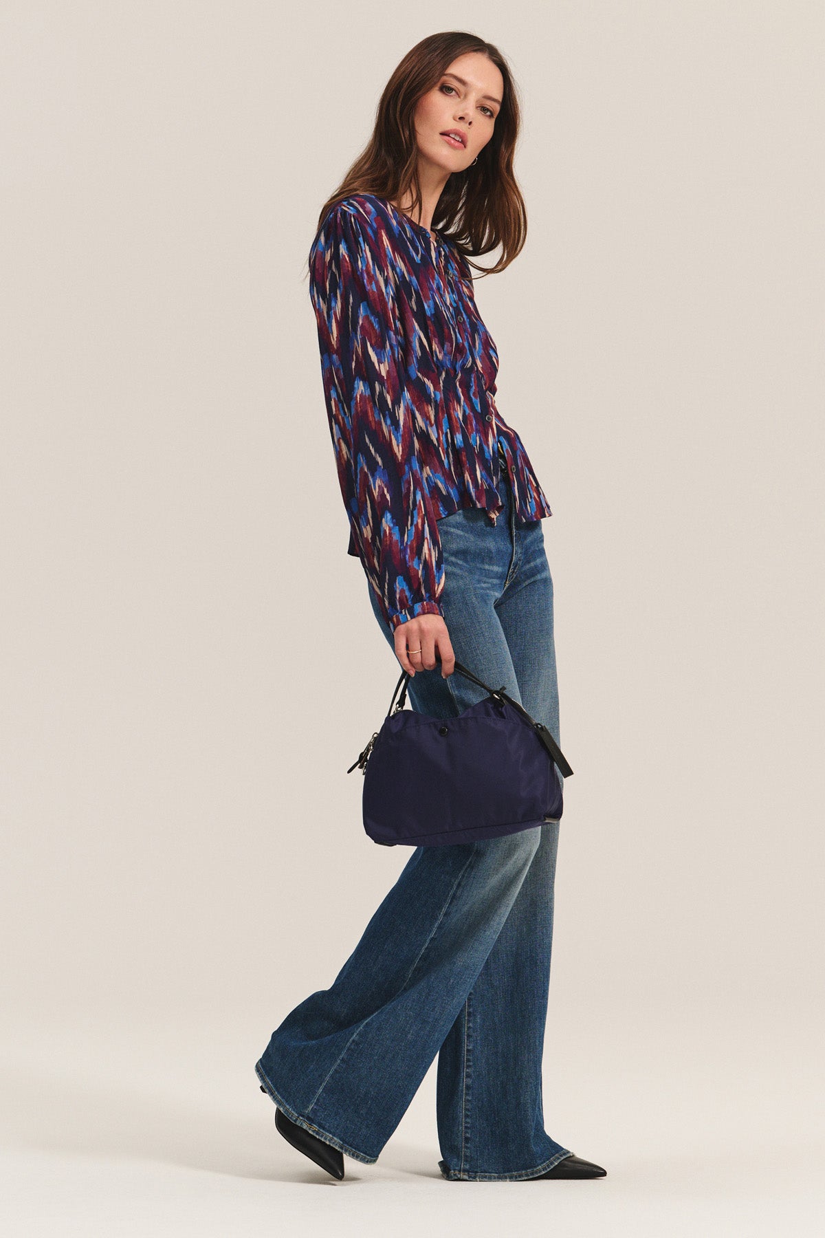 A woman wears the ESTELLA BLOUSE by Velvet by Graham & Spencer—a colorful cotton viscose top—paired with flared blue jeans, black pointed shoes, and a dark blue handbag against a plain light background.-40732534440129