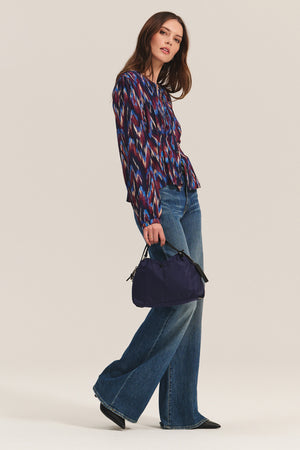A woman wears the ESTELLA BLOUSE by Velvet by Graham & Spencer—a colorful cotton viscose top—paired with flared blue jeans, black pointed shoes, and a dark blue handbag against a plain light background.