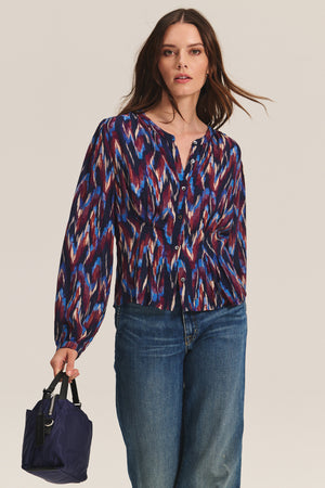 A woman models the ESTELLA BLOUSE by Velvet by Graham & Spencer—a multicolored, long-sleeve cotton-viscose top—paired with blue jeans and a small navy handbag against a plain background.