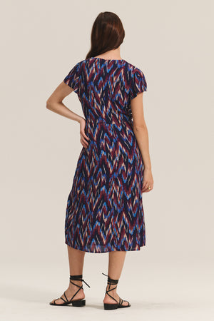 A woman stands facing away, wearing the MIRANDA DRESS by Velvet by Graham & Spencer, a cotton-viscose midi with a blue, red, and white abstract print and black strappy sandals that highlight her feminine silhouette.
