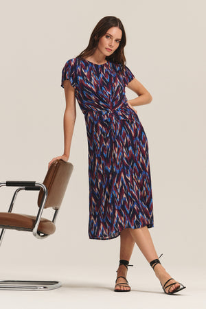 A woman wearing the MIRANDA DRESS by Velvet by Graham & Spencer, a colorful patterned cotton-viscose midi, stands beside a brown chair with one arm resting on it and the other on her hip, paired with black strappy sandals.