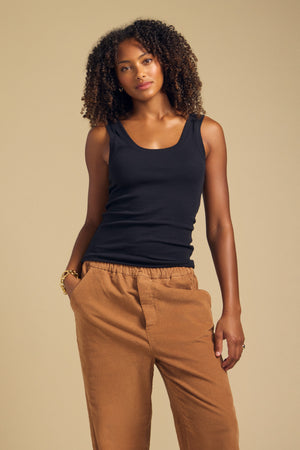 A woman with curly hair wears the MOLLY TANK TOP by Velvet by Graham & Spencer in black, paired with tan pants and standing with one hand in her pocket against a beige background.