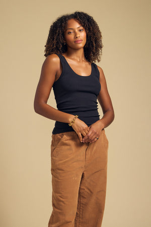 A woman with curly hair stands against a beige background, wearing the Velvet by Graham & Spencer MOLLY TANK TOP in black and brown pants, with her hands clasped in front of her.