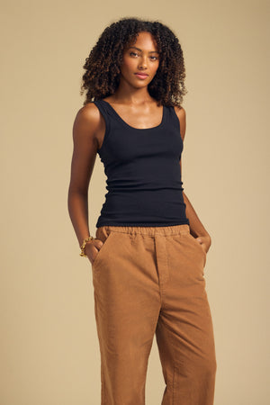 A person stands against a plain beige background, wearing the MOLLY TANK TOP by Velvet by Graham & Spencer in black, paired with tan wide-leg pants. Their hands are in their pockets and they have a neutral expression.