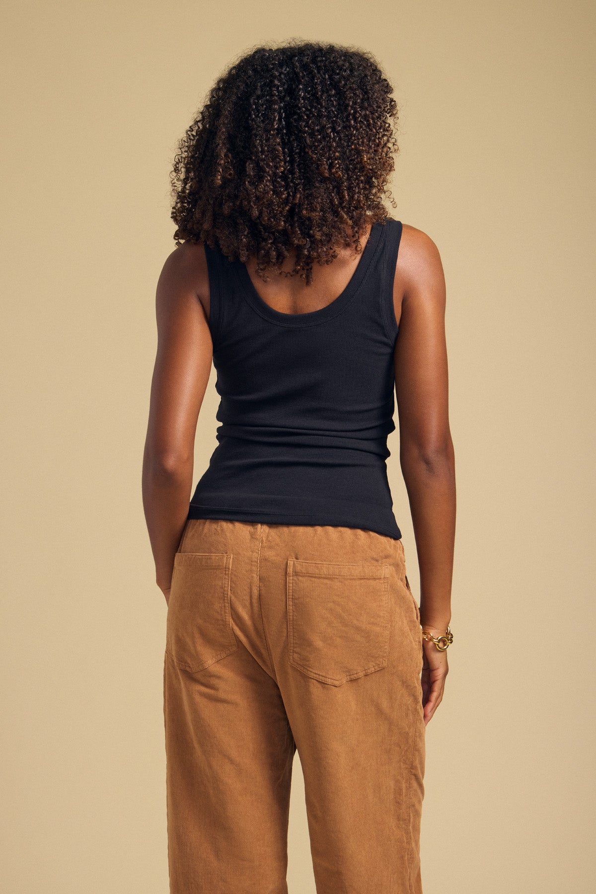 Wearing the Velvet by Graham & Spencer MOLLY TANK TOP in black, a person with curly hair and tan pants stands facing away against a plain beige background.