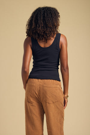 Wearing the Velvet by Graham & Spencer MOLLY TANK TOP in black, a person with curly hair and tan pants stands facing away against a plain beige background.