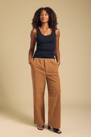 A woman stands against a beige background wearing the Velvet by Graham & Spencer JAYDA CORDUROY PANT—a cotton corduroy wide-leg style with a drawstring elastic waist—paired with a black sleeveless top, black open-toe heels, and her hands in her pockets.