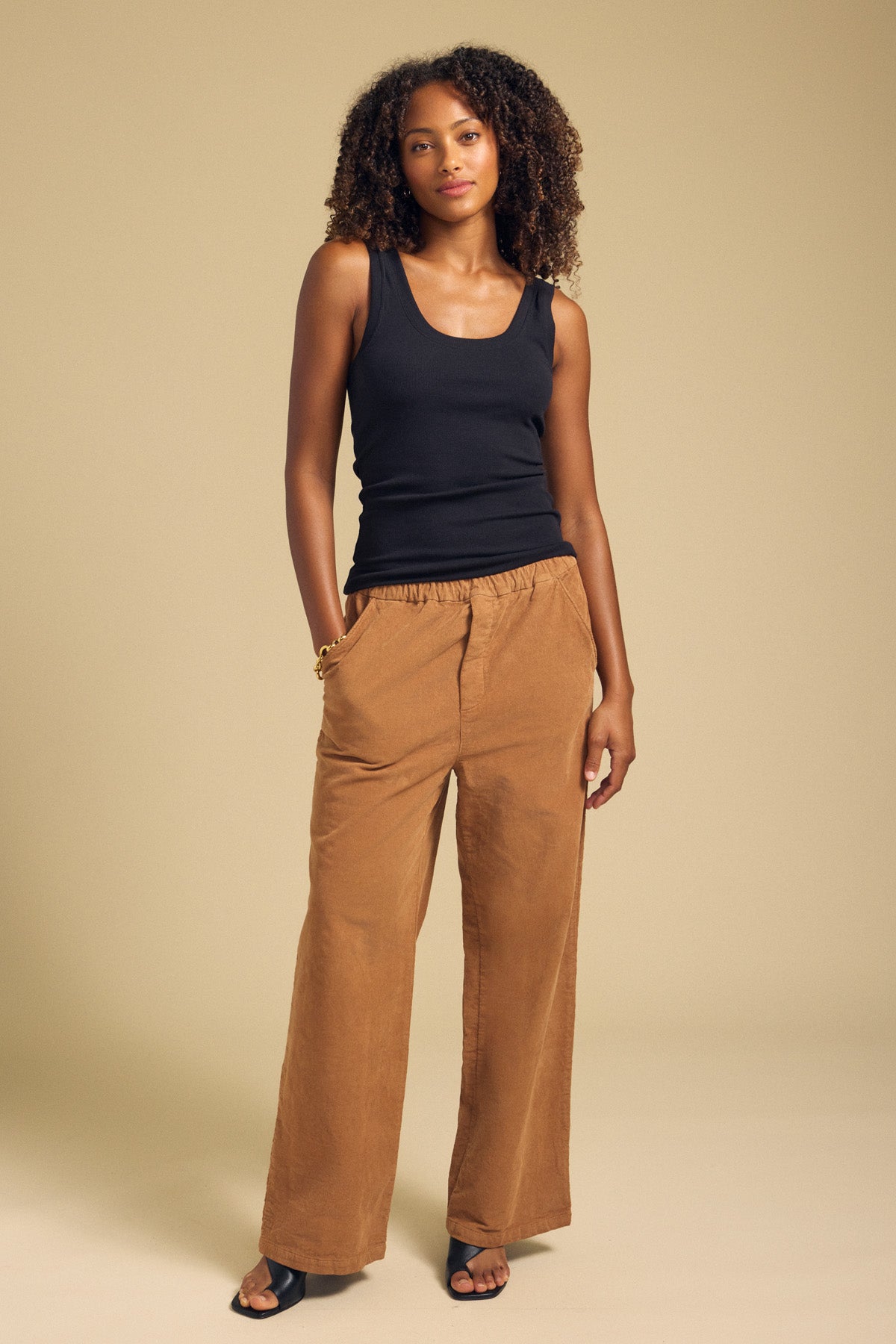 A woman stands against a plain beige background with her hands in the pockets of the Velvet by Graham & Spencer JAYDA CORDUROY PANT—a pair of tan cotton corduroy wide-leg pants featuring a drawstring elastic waist. She pairs them with a black tank top and black heeled sandals.
