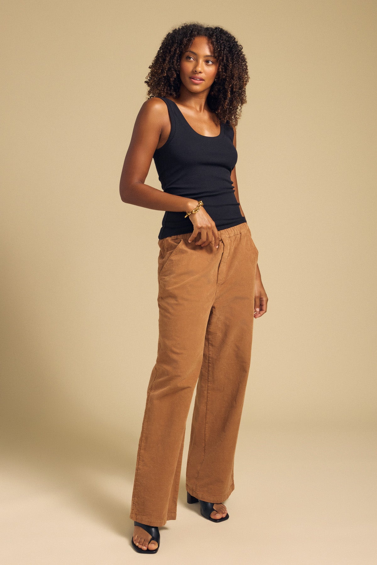 A woman stands on a neutral background wearing a black sleeveless top, the JAYDA CORDUROY PANT by Velvet by Graham & Spencer in brown with a wide-leg silhouette and drawstring elastic waist, paired with black heeled sandals.