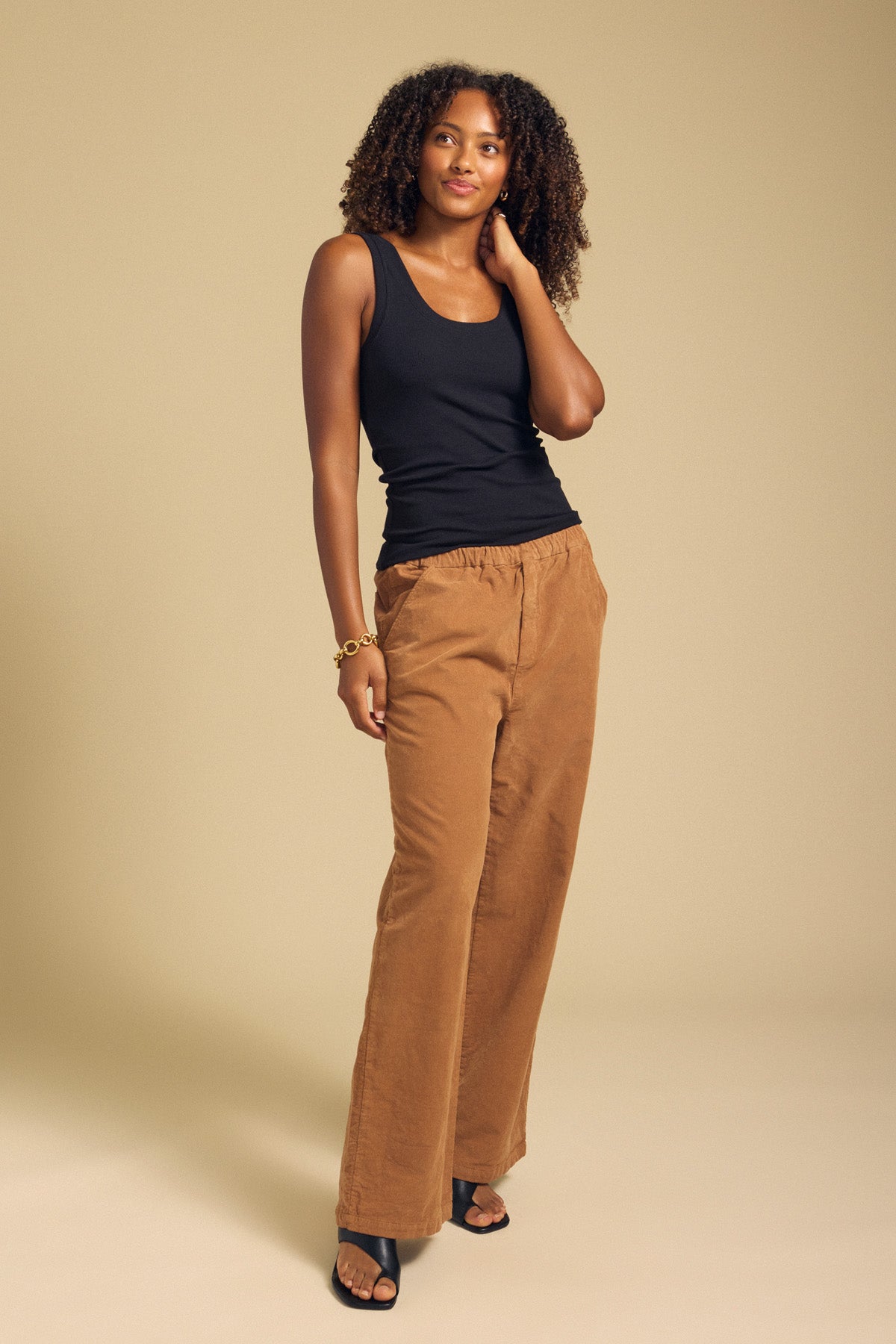 A woman stands against a beige background wearing the MOLLY TANK TOP by Velvet by Graham & Spencer—a black ribbed tank featuring a fitted silhouette—paired with brown wide-leg pants, black sandals, and a bracelet, with one hand touching her hair.