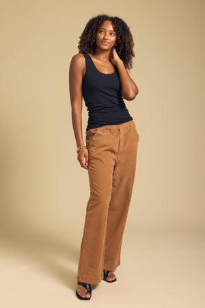 A woman stands against a beige background wearing the MOLLY TANK TOP by Velvet by Graham & Spencer—a black ribbed tank featuring a fitted silhouette—paired with brown wide-leg pants, black sandals, and a bracelet, with one hand touching her hair.
