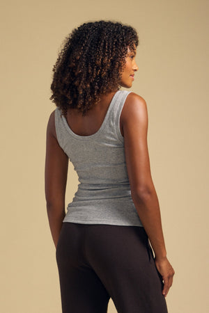 A person with curly hair is shown from the back against a plain beige background, wearing the MOLLY TANK TOP by Velvet by Graham & Spencer in gray and dark pants. The fitted silhouette of this modal knit tank top lends a touch of modern simplicity to the look.