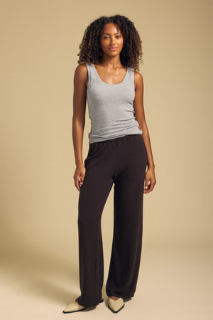 A woman stands against a beige background wearing the MOLLY TANK TOP in gray modal ribbed knit by Velvet by Graham & Spencer, paired with loose black pants and cream pointed shoes.