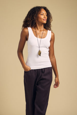 A person with curly hair wears the MOLLY TANK TOP by Velvet by Graham & Spencer—a white ribbed tank top featuring a fitted silhouette—paired with dark pants and a long necklace, standing with one hand in their pocket against a plain beige background.