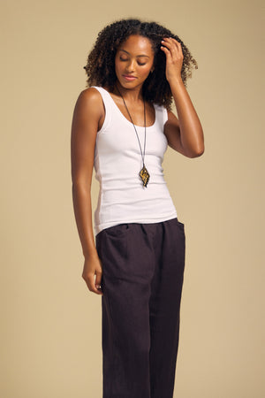 A woman wears the MOLLY TANK TOP by Velvet by Graham & Spencer—a white ribbed, fitted silhouette—paired with dark pants. She stands against a beige background, touching her hair with one hand and looking down.
