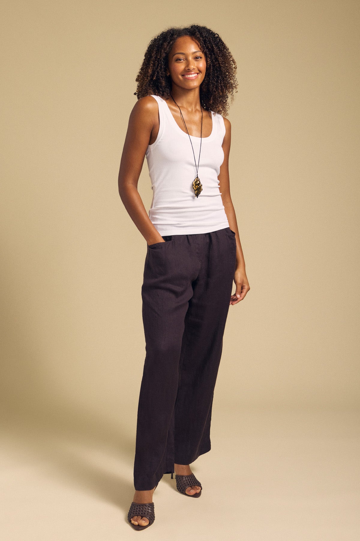 A woman stands smiling against a plain beige background, wearing the MOLLY TANK TOP by Velvet by Graham & Spencer—a white ribbed tank featuring a fitted silhouette—paired with black wide-leg pants, a long pendant necklace, and open-toed shoes.