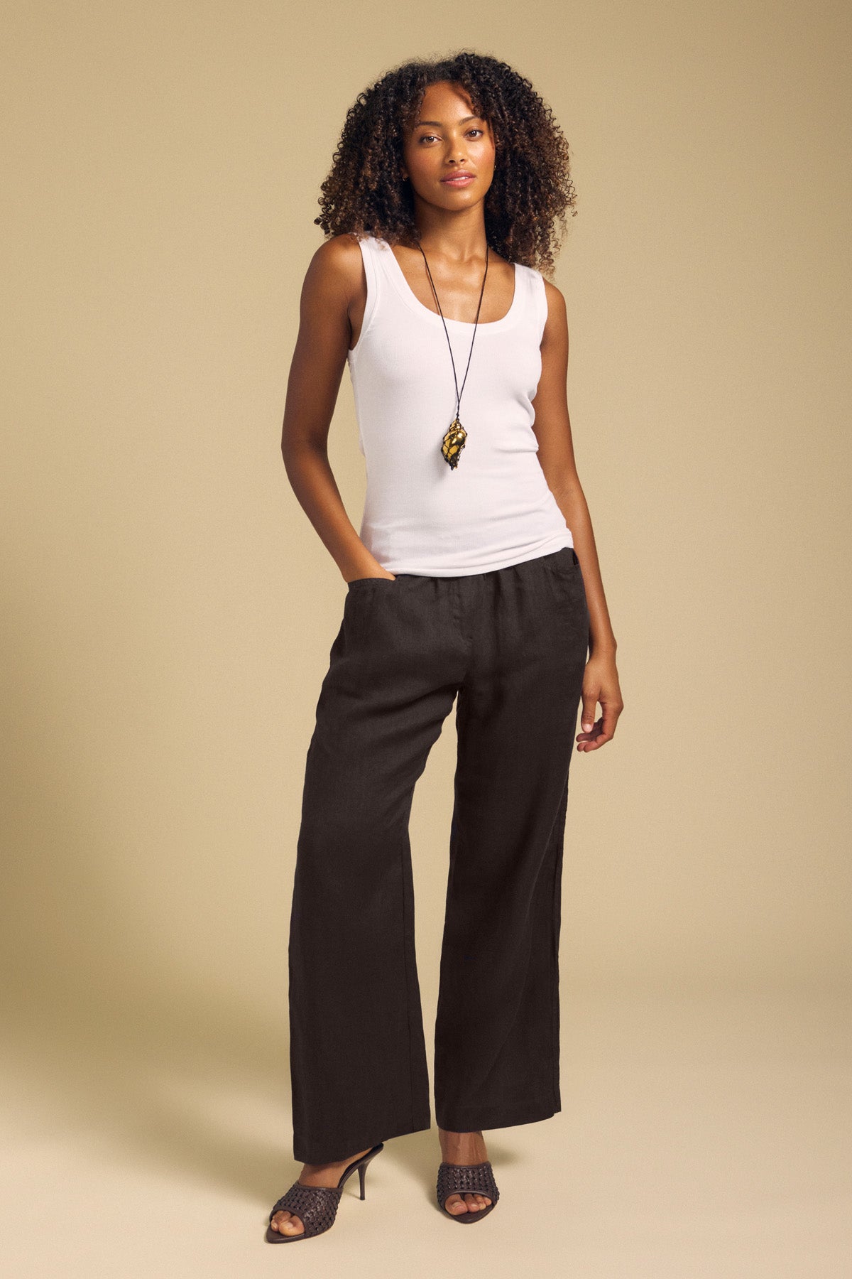 A woman stands against a beige background wearing the Velvet by Graham & Spencer CLAIRO LINEN PANT in black, styled with a white sleeveless top, black heels, and a long pendant necklace.