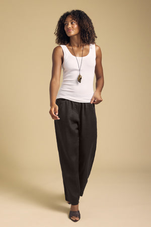 A woman stands against a beige background wearing a white sleeveless top, black CLAIRO LINEN PANT by Velvet by Graham & Spencer—featuring wide legs and an elastic waist—paired with black sandals and a long necklace.