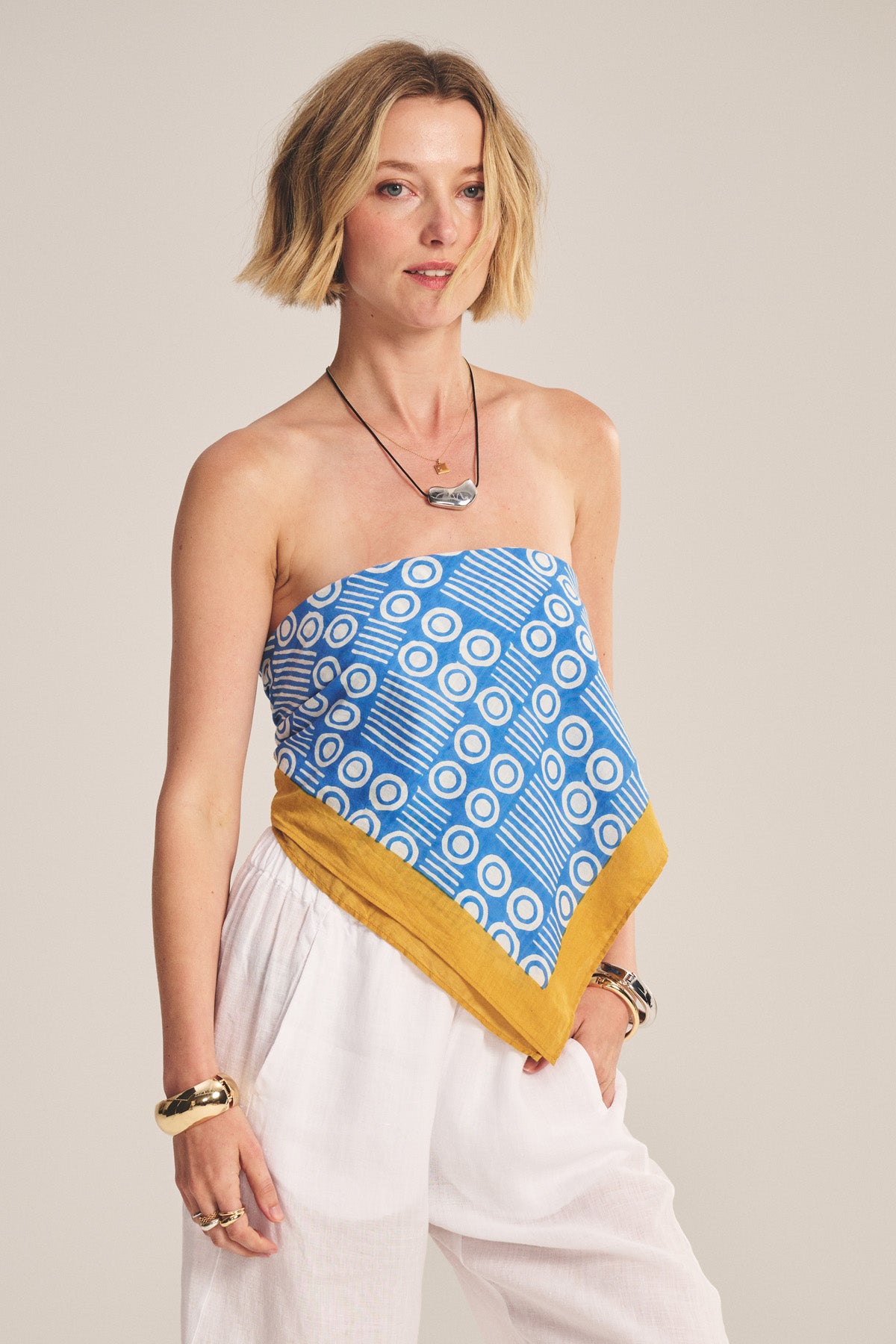 A woman in white pants wears the MONROE SCARF by Velvet by Graham & Spencer—a blue geometric patterned, lightweight cotton scarf top with a yellow border—styled with bracelets and necklaces against a neutral background. A versatile accessory.-40808673804481