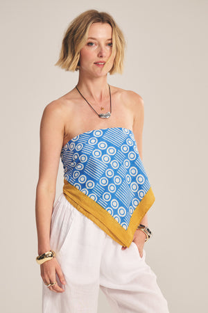 A woman in white pants wears the MONROE SCARF by Velvet by Graham & Spencer—a blue geometric patterned, lightweight cotton scarf top with a yellow border—styled with bracelets and necklaces against a neutral background. A versatile accessory.