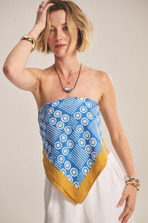 A woman styles the MONROE SCARF by Velvet by Graham & Spencer—a versatile, lightweight cotton scarf—over a strapless blue and white geometric top with a yellow border, white pants, and jewelry, posed against a plain background.