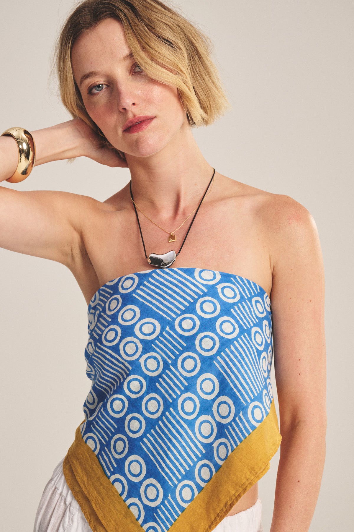 A woman with short blonde hair poses with one hand behind her neck, wearing the MONROE SCARF by Velvet by Graham & Spencer as a blue geometric-patterned versatile top in lightweight cotton, paired with gold jewelry against a plain background.-40808673673409