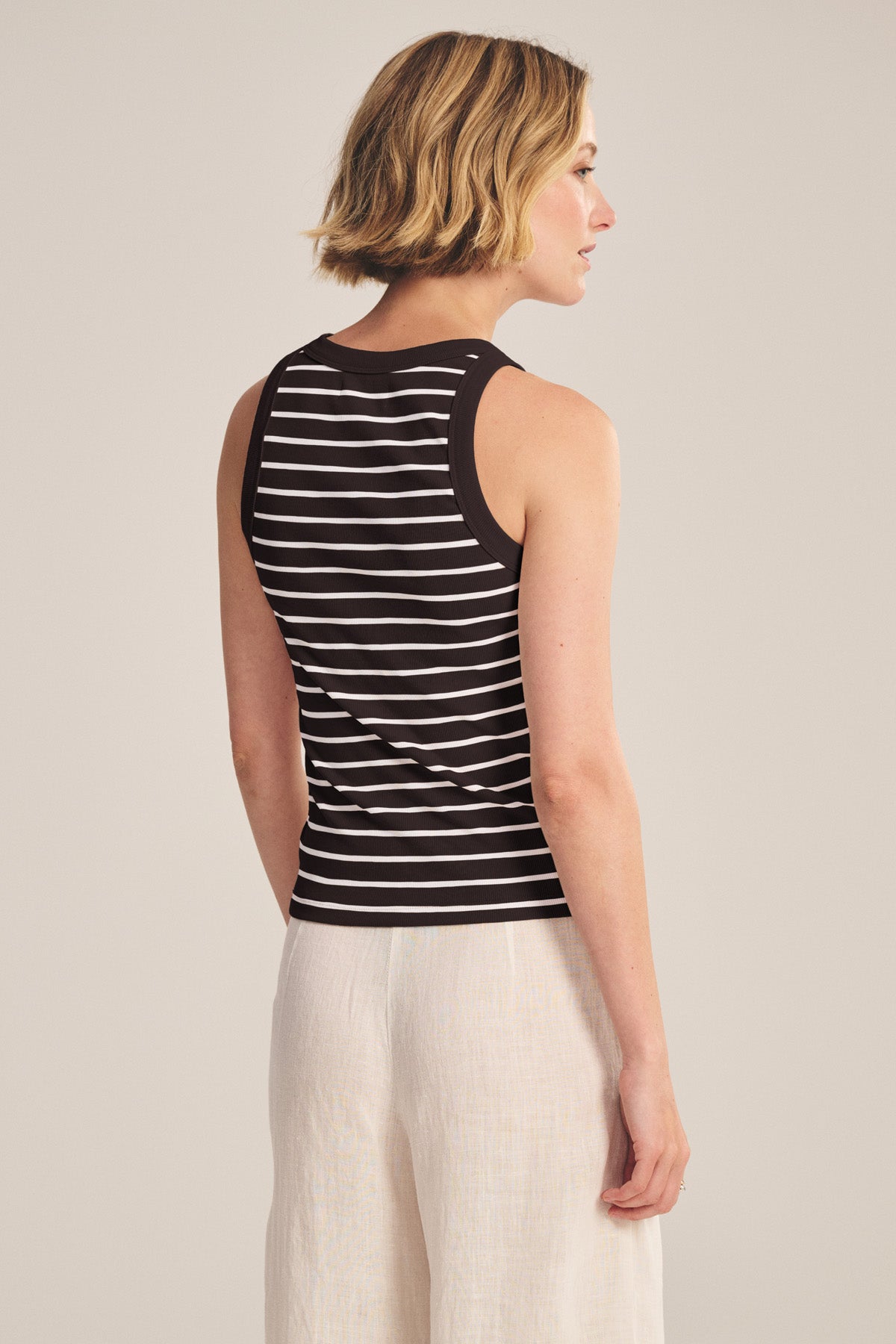 A woman stands facing away, wearing the MORIE TANK TOP by Velvet by Graham & Spencer—a striped cotton tank—paired with light-colored pants against a plain background.-41044594360513