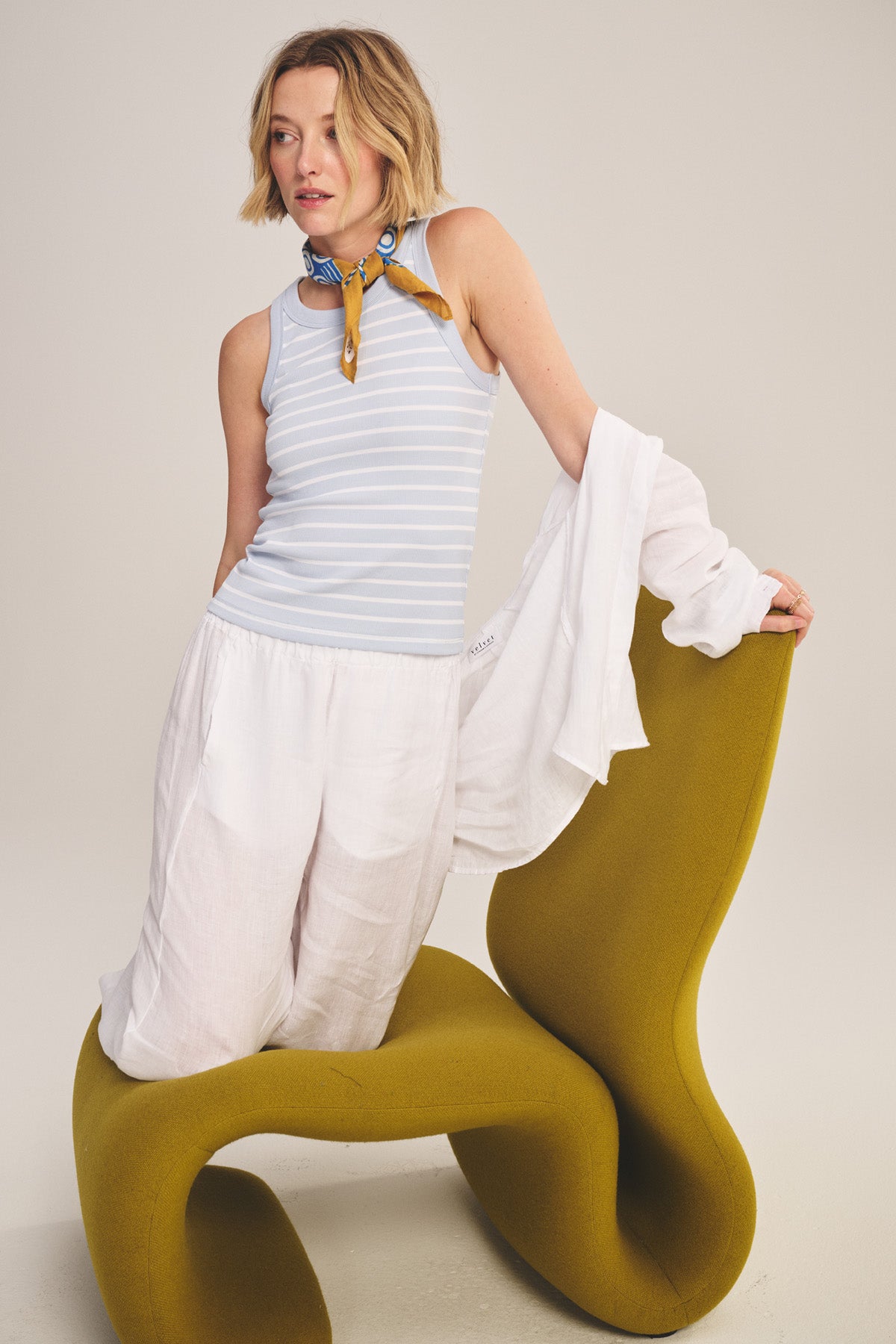 A woman wearing the MORIE TANK TOP by Velvet by Graham & Spencer, styled with white pants and a yellow scarf, poses on a modern green chair while holding a white shirt.-41044015415489